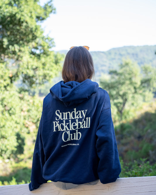 Person wearing a blue hoodie with 'Sunday Pickleball Club' text, sitting outdoors with greenery and mountains in the background.